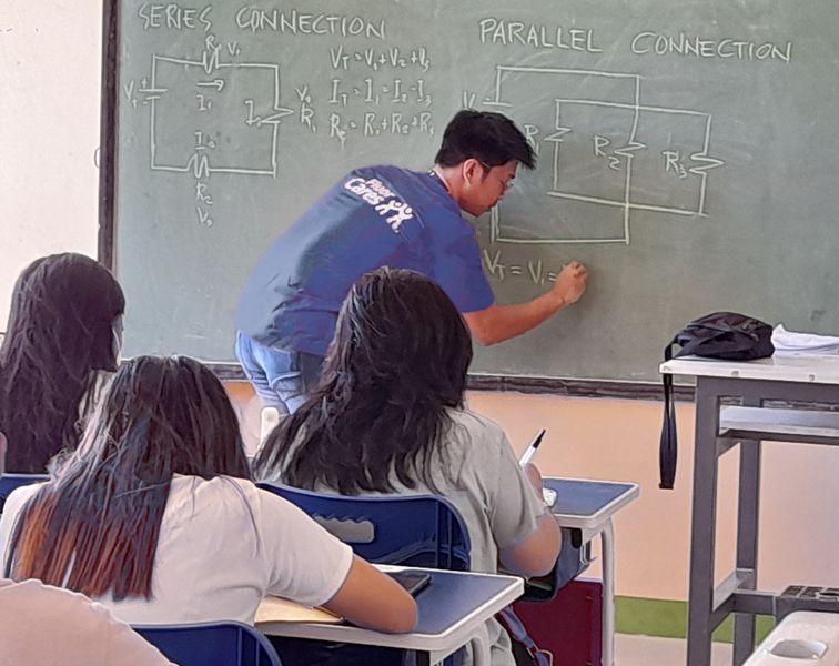 Fluor volunteer drawing circuits on a blackboard in front of a class with three seated girls shown